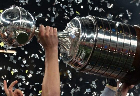 Close-up da taça da Libertadores da América, mostrando os detalhes em prata e a base de madeira com as placas dos clubes campeões.