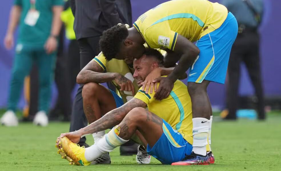 Atletas com a camisa amarela da Seleção Brasileira consolam Neymar sentado no gramado chorando e cabisbaixo após o apito final na Copa de 2026.